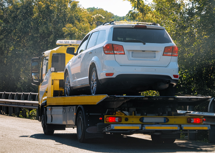 Una grúa de plataforma amarilla y negra transporta un automóvil blanco por la carretera. El vehículo está asegurado firmemente sobre la plataforma, reflejando un escenario común de remolque, ya sea por avería o traslado programado. En el fondo, árboles y vegetación enmarcan la escena, ofreciendo un contraste entre el tráfico y el entorno natural.