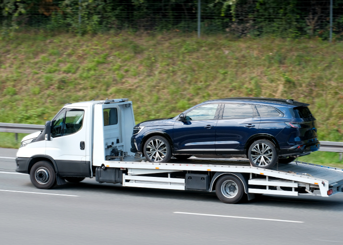 Una grúa blanca transporta un SUV azul oscuro por una carretera. El vehículo está asegurado sobre la plataforma de la grúa, que se encuentra en movimiento. La imagen muestra un proceso de remolque, un servicio esencial en situaciones de emergencia o traslado de vehículos.