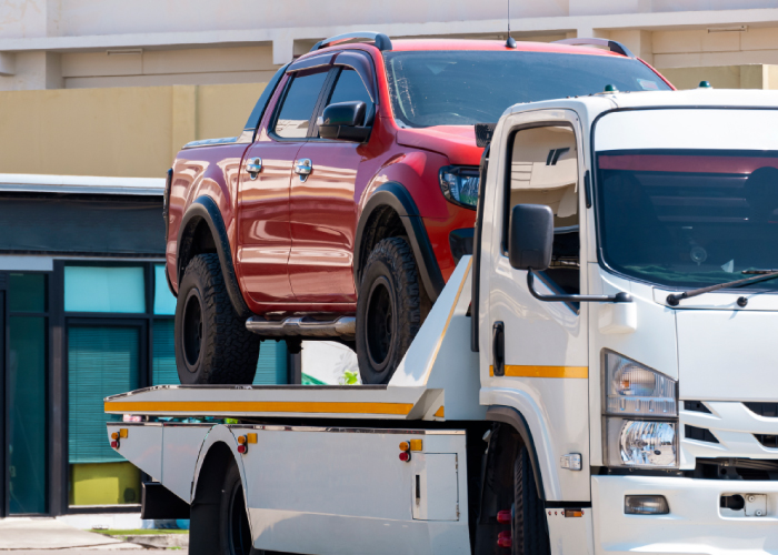 Una camioneta roja está asegurada sobre la plataforma de una grúa blanca en movimiento. La escena ocurre en un entorno urbano con edificios visibles al fondo, representando el transporte de vehículos por avería, accidente o traslado.