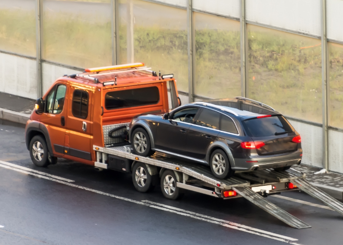Una camioneta de remolque naranja transporta un automóvil gris oscuro sobre una plataforma. La camioneta tiene luces de emergencia en el techo y rampas extendidas en la parte trasera para facilitar la carga y descarga del vehículo. La escena ocurre en una carretera de dos carriles con una barrera de vidrio al fondo, destacando el proceso de traslado de vehículos.
