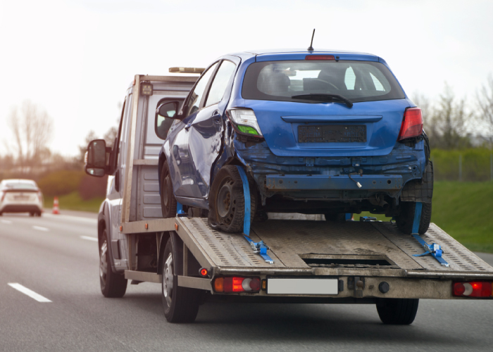 Grúa de remolque transportando un automóvil azul con daños en la parte trasera, asegurado con correas azules sobre la plataforma, en movimiento por una carretera.