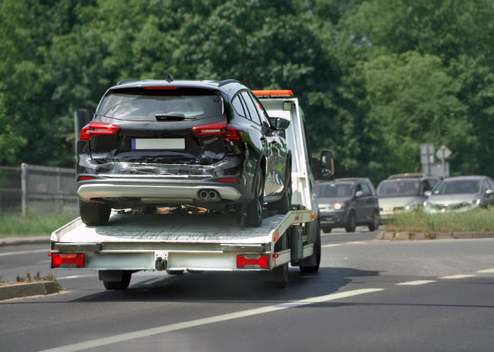 Automóvil negro con daños en la parte trasera, transportado en una grúa sobre una carretera. La parte trasera del vehículo está abollada y rasgada, indicando un posible accidente. En el fondo se ven otros vehículos y árboles.