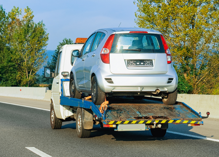Camión de remolque transportando un vehículo plateado por una carretera, con árboles y cielo azul de fondo.