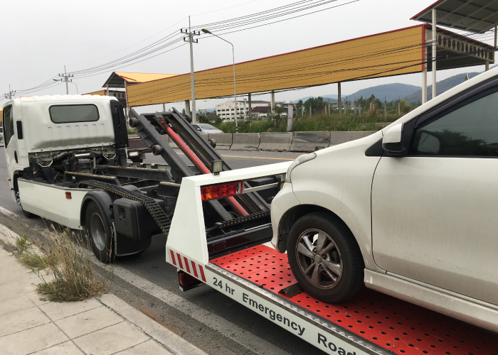 Una grúa roja con franjas blancas transporta un automóvil blanco asegurado en su plataforma. La grúa tiene un letrero que indica servicio de emergencia 24 horas en carretera. Está estacionada junto a una acera, con postes de electricidad y una estructura de techo amarillo en el fondo. La escena refleja un servicio de asistencia vehicular en acción.