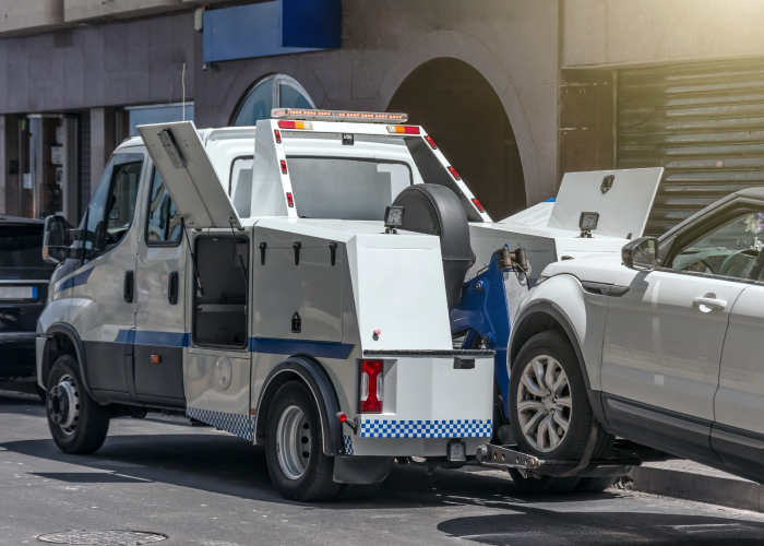 Una grúa blanca con detalles azules remolca un automóvil blanco en una calle urbana. La grúa tiene varias puertas abiertas que muestran compartimentos de almacenamiento. En el fondo, se ven edificios y otros vehículos, situando la escena en un entorno citadino.