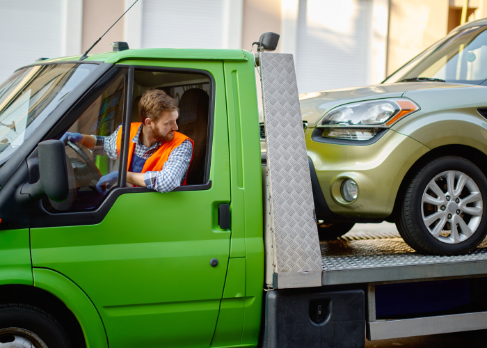 Un camión de remolque verde transporta un automóvil dorado. El conductor, con chaleco de seguridad naranja, observa desde la cabina. El vehículo dorado está asegurado sobre la plataforma, listo para ser trasladado.