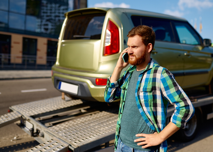 Una persona de pie junto a una grúa de remolque que transporta un vehículo verde, mientras habla por teléfono móvil. La persona viste una camisa de cuadros con una camiseta verde debajo. La escena ocurre en una calle urbana con edificios en el fondo.