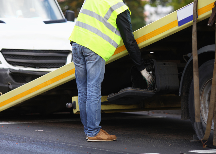 Una persona con chaleco reflectante amarillo y jeans azules trabaja en la parte trasera de un camión de remolque, manipulando una caja o compartimento. La rampa inclinada del camión sugiere una tarea relacionada con el transporte o mantenimiento de vehículos en una vía pública.