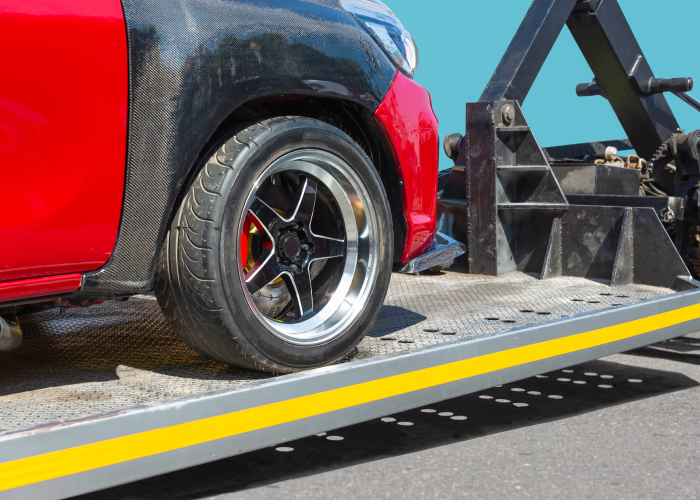 Un automóvil rojo con una llanta de aleación negra y plateada está siendo cargado en una plataforma de remolque metálica perforada con un borde amarillo. La estructura negra del remolque asegura el vehículo durante el transporte, mostrando un proceso típico de remolque.